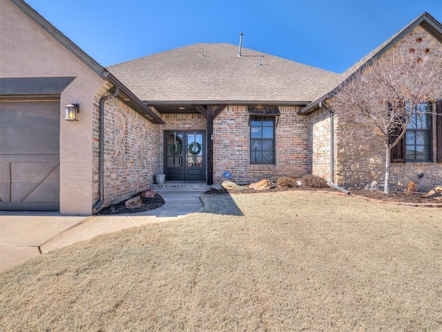 doorway to property with a shingled roof, french doors, an attached garage, a yard, and brick siding