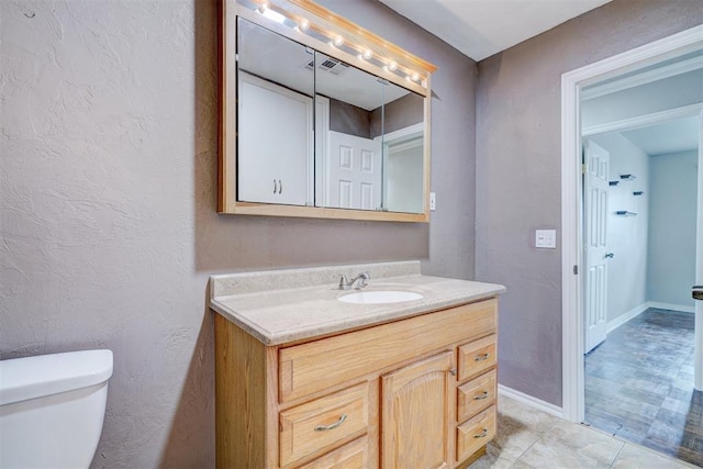 half bathroom featuring vanity, visible vents, baseboards, toilet, and a textured wall