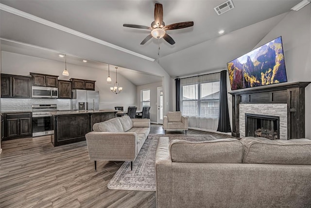 living area featuring visible vents, vaulted ceiling, a stone fireplace, wood finished floors, and ceiling fan with notable chandelier