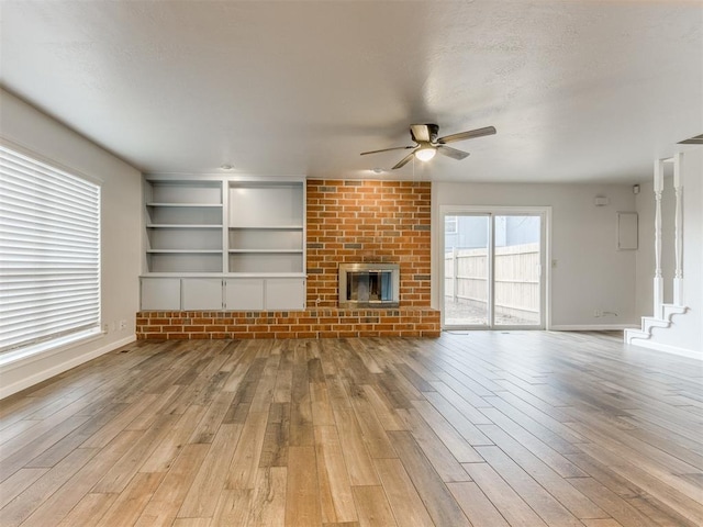 unfurnished living room featuring a ceiling fan, built in features, baseboards, light wood-type flooring, and a brick fireplace