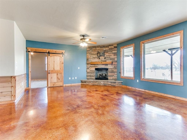 unfurnished living room featuring visible vents, a barn door, ceiling fan, a stone fireplace, and baseboards