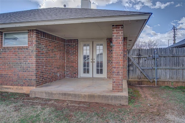 entrance to property featuring brick siding, a shingled roof, fence, and french doors