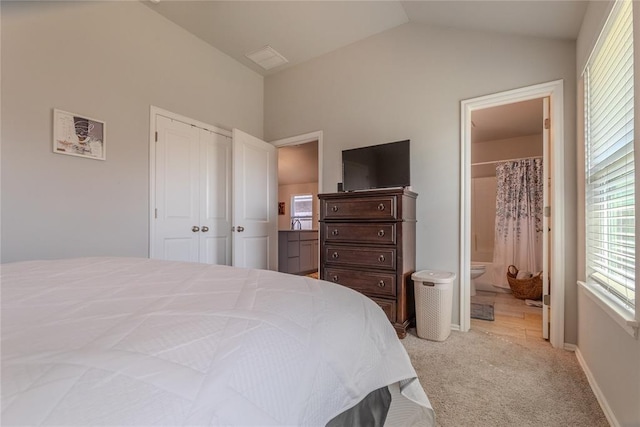 bedroom featuring a closet, light colored carpet, vaulted ceiling, ensuite bath, and baseboards