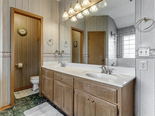 bathroom featuring double vanity, a sink, toilet, and wallpapered walls