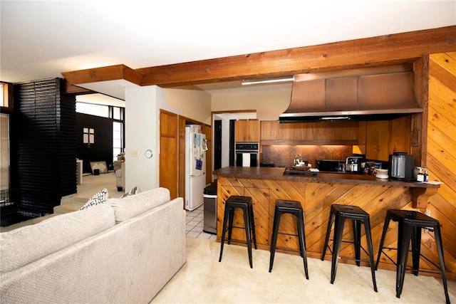 kitchen with oven, wall chimney range hood, freestanding refrigerator, brown cabinetry, and dark countertops