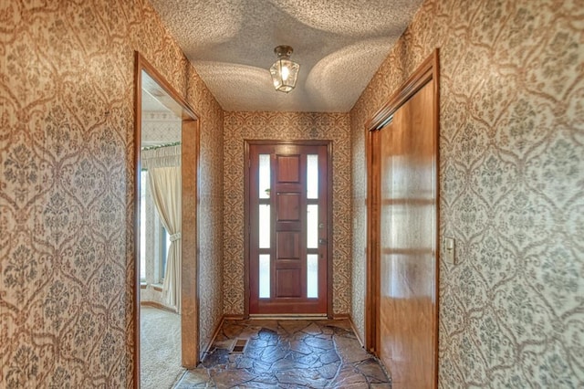 doorway to outside with stone tile floors, a textured ceiling, wallpapered walls, and baseboards