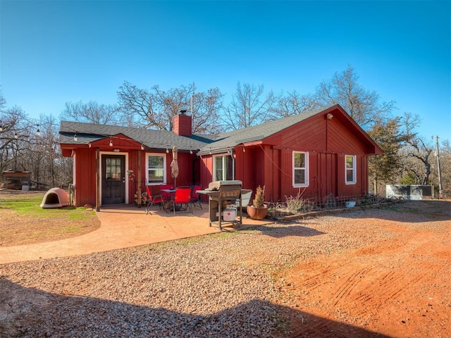 rear view of house featuring board and batten siding, a patio area, a shingled roof, and a chimney