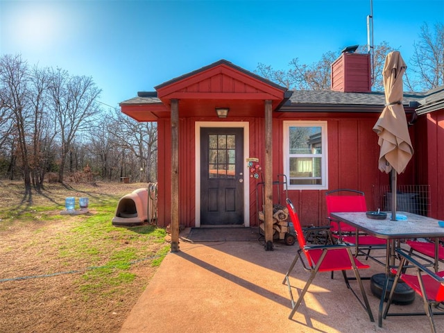 view of exterior entry featuring roof with shingles, a patio, a chimney, a lawn, and board and batten siding