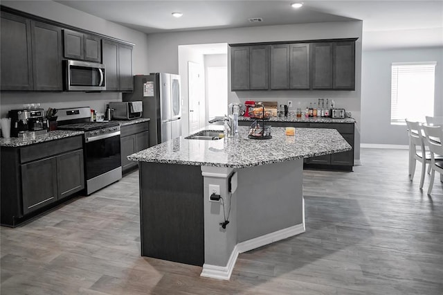 kitchen with light stone counters, appliances with stainless steel finishes, a kitchen island with sink, a sink, and baseboards