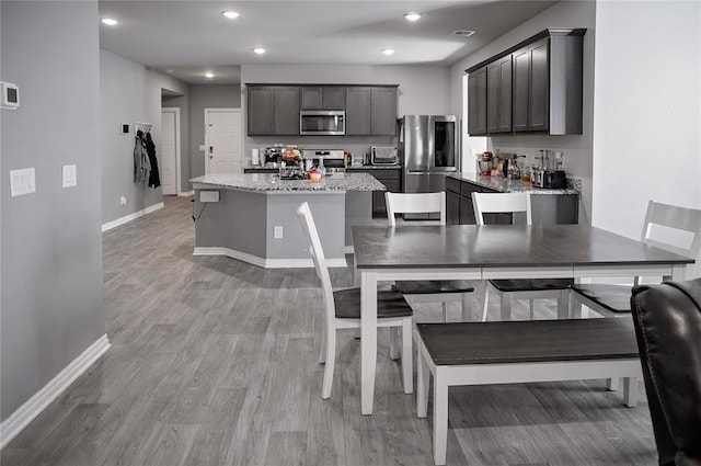 kitchen featuring stainless steel appliances, a kitchen island with sink, light stone countertops, light wood-type flooring, and baseboards