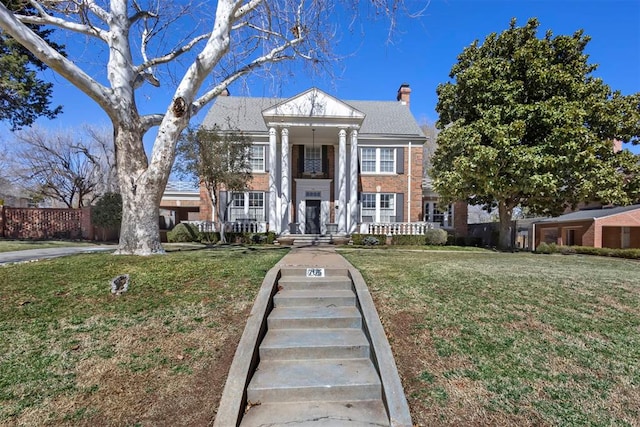 neoclassical home with a front yard, covered porch, brick siding, and a chimney