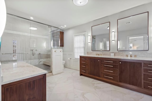 bathroom featuring marble finish floor, a wainscoted wall, double vanity, toilet, and a stall shower