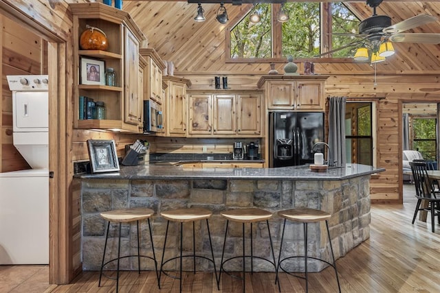 kitchen featuring wooden walls, stainless steel microwave, stacked washer / drying machine, and black fridge with ice dispenser