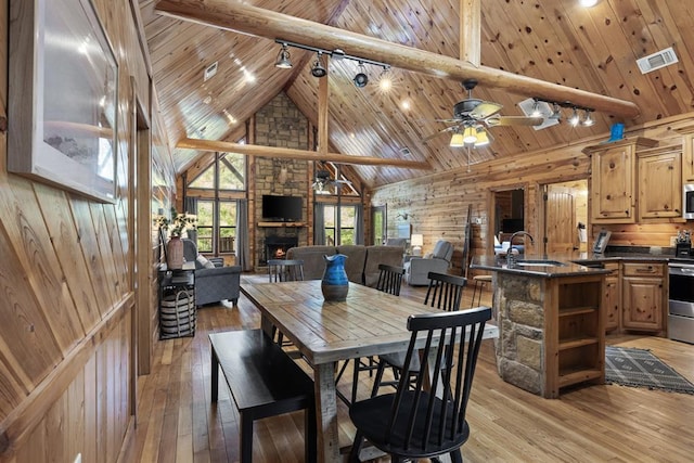 dining room featuring wooden walls, wood ceiling, beamed ceiling, a stone fireplace, and light wood-style floors