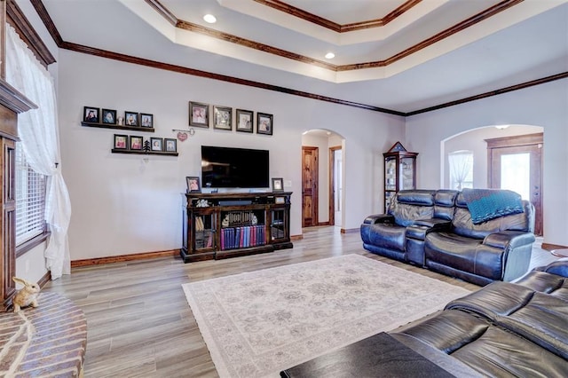 living room featuring baseboards, arched walkways, ornamental molding, a tray ceiling, and light wood-style floors