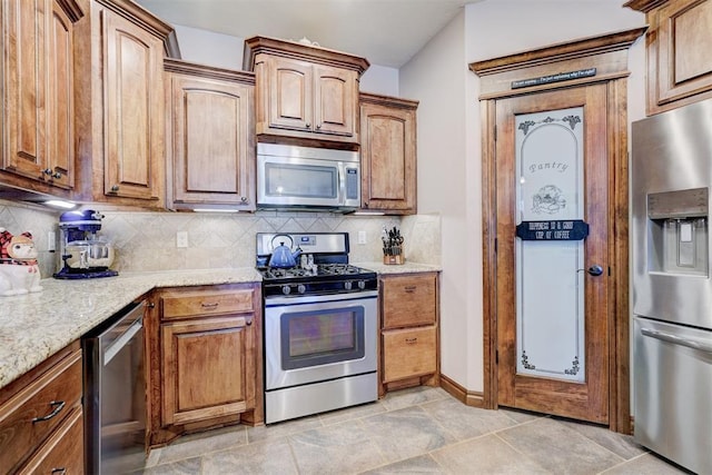 kitchen featuring decorative backsplash, appliances with stainless steel finishes, brown cabinetry, light stone countertops, and baseboards