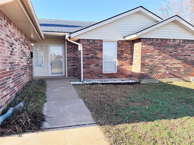 doorway to property with a yard, brick siding, and roof with shingles