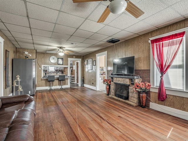 unfurnished living room with wooden walls, a drop ceiling, ceiling fan, wood-type flooring, and a fireplace