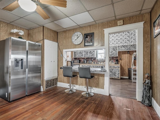 kitchen with a breakfast bar area, a drop ceiling, wood walls, wood finished floors, and stainless steel fridge