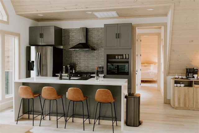 kitchen with wall chimney exhaust hood, a kitchen bar, gray cabinets, and stainless steel fridge with ice dispenser