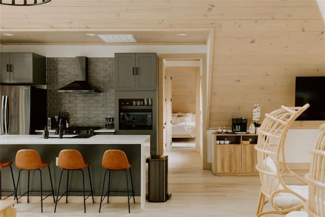 kitchen with wall oven, light wood finished floors, wall chimney exhaust hood, a kitchen breakfast bar, and light countertops