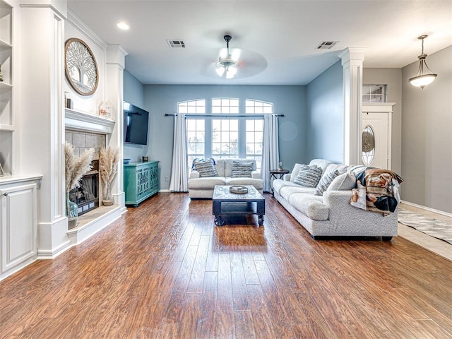 living area featuring visible vents, a ceiling fan, a tile fireplace, wood finished floors, and ornate columns