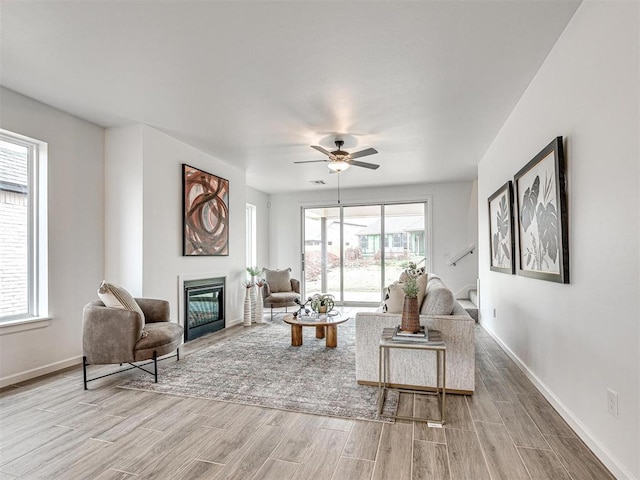 living room featuring a glass covered fireplace, ceiling fan, light wood-style flooring, and baseboards