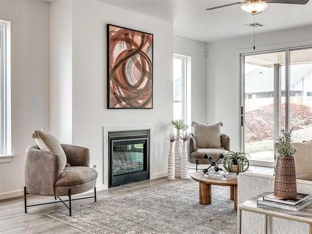 living area featuring visible vents, baseboards, a glass covered fireplace, ceiling fan, and light wood-type flooring