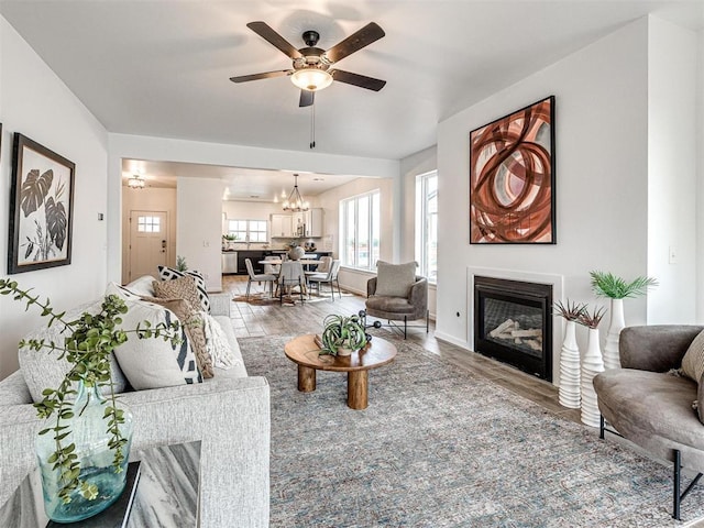 living room featuring light wood-style floors, a glass covered fireplace, baseboards, and ceiling fan with notable chandelier