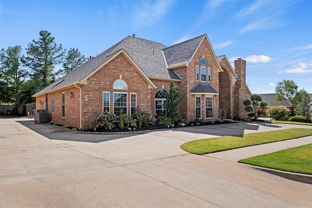 view of front of property with central AC unit, brick siding, driveway, roof with shingles, and a chimney