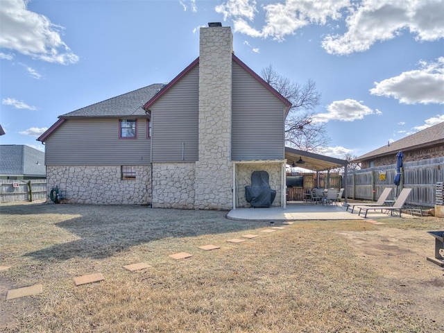 rear view of house featuring roof with shingles, a patio, a chimney, fence, and stone siding