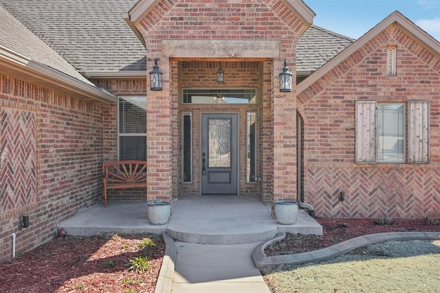 doorway to property with covered porch, brick siding, and roof with shingles