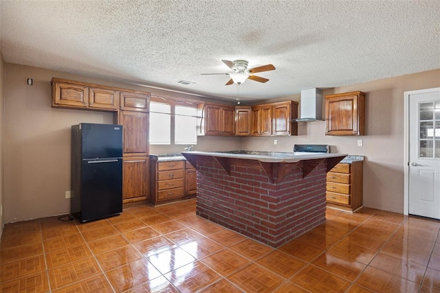 kitchen featuring range, wall chimney exhaust hood, brown cabinets, freestanding refrigerator, and light countertops