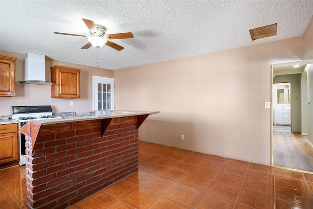 kitchen featuring wall chimney range hood, white range with gas cooktop, brown cabinetry, and light countertops