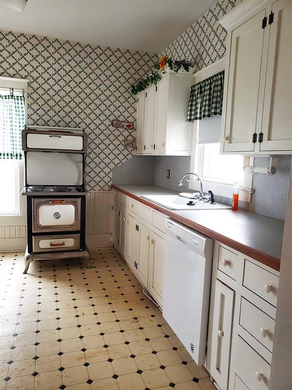 kitchen featuring light floors, white cabinetry, white dishwasher, a sink, and wallpapered walls