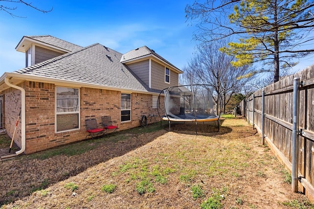 rear view of house featuring a trampoline, a fenced backyard, a yard, roof with shingles, and brick siding