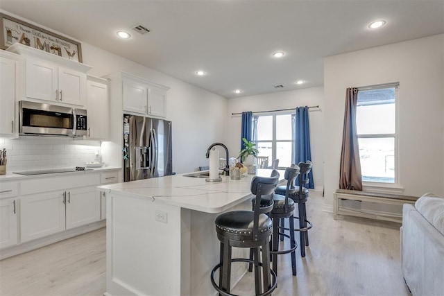 kitchen with a kitchen island with sink, a sink, appliances with stainless steel finishes, light wood-type flooring, and decorative backsplash