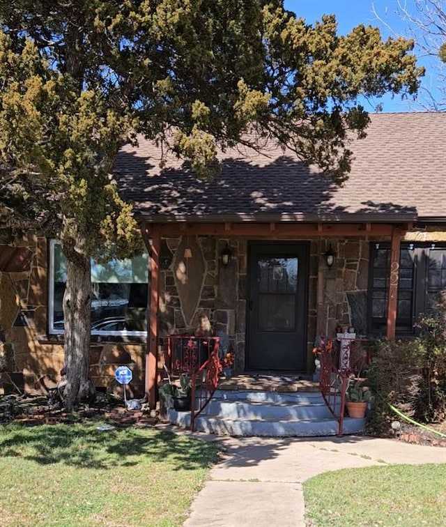 doorway to property featuring stone siding, a lawn, roof with shingles, and covered porch