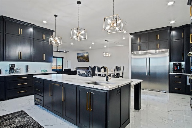 kitchen with an island with sink, dark cabinets, stainless steel built in fridge, marble finish floor, and a sink