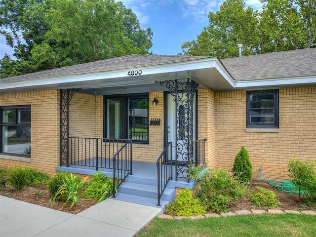 property entrance featuring a porch, roof with shingles, and brick siding