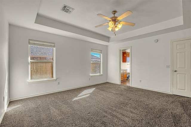 carpeted empty room with a tray ceiling, a ceiling fan, visible vents, and baseboards