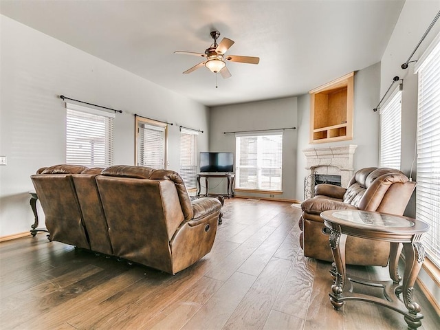 living area with a wealth of natural light, ceiling fan, wood finished floors, and a stone fireplace