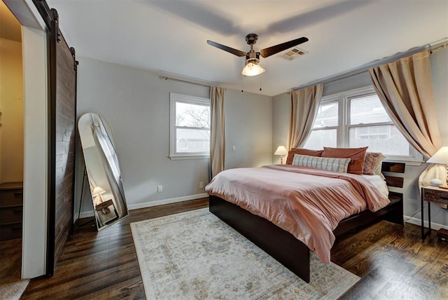 bedroom with ceiling fan, a barn door, visible vents, baseboards, and dark wood finished floors