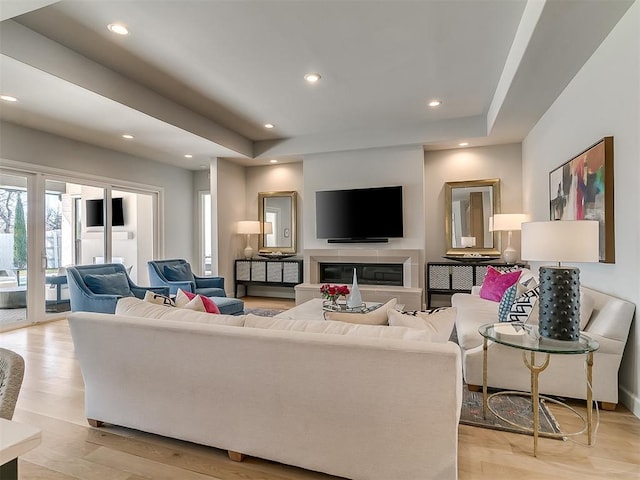 living room with recessed lighting, a tray ceiling, a glass covered fireplace, and light wood-style flooring