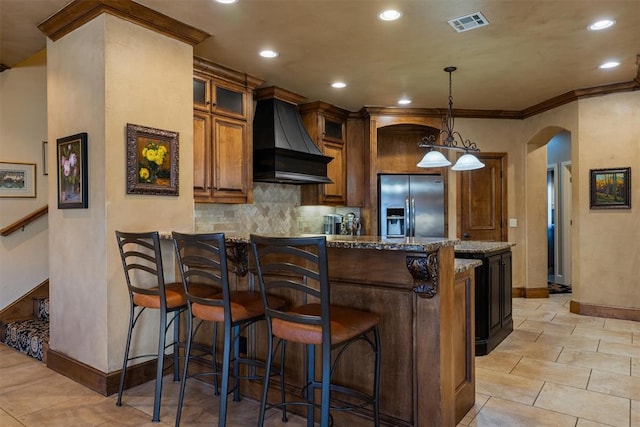 kitchen with arched walkways, stone countertops, stainless steel fridge with ice dispenser, a kitchen bar, and custom range hood