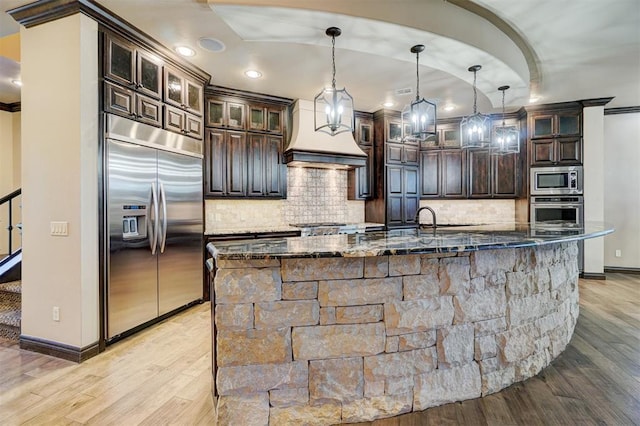 kitchen featuring dark stone counters, built in appliances, dark brown cabinets, premium range hood, and backsplash