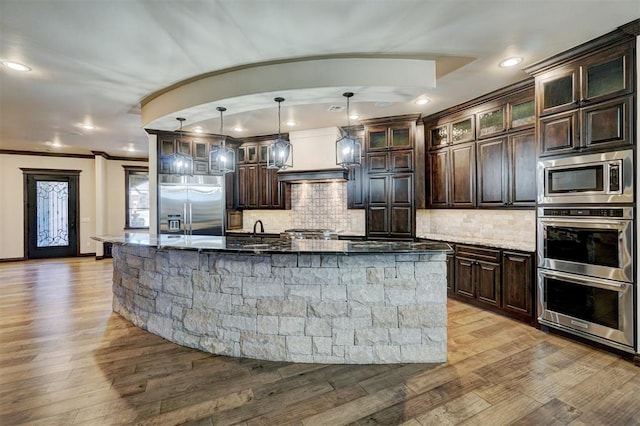 kitchen with a large island, decorative backsplash, dark brown cabinetry, wood finished floors, and built in appliances