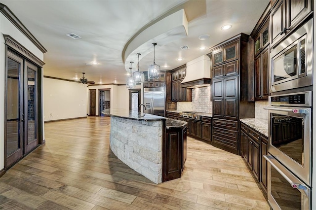 kitchen featuring custom range hood, visible vents, dark brown cabinets, built in appliances, and dark stone counters