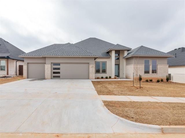 prairie-style home with an attached garage, a shingled roof, concrete driveway, and brick siding