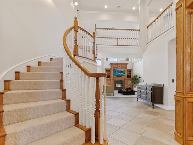 staircase with crown molding, recessed lighting, a towering ceiling, a glass covered fireplace, and tile patterned flooring